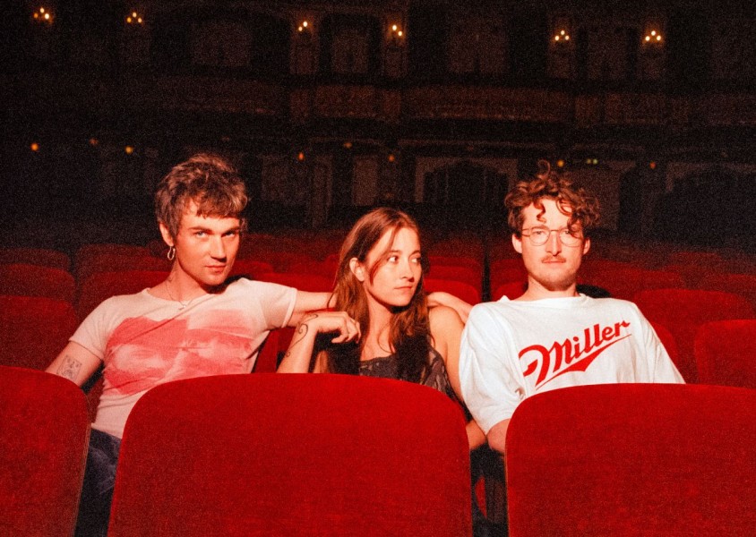 Three people sit in red theater seats, with the two on the sides leaning toward the person in the middle.x